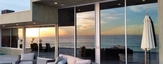 A modern patio featuring woven outdoor furniture, including a sofa and chairs, facing the ocean. A large glass wall reflects a picturesque sunset over the sea, with outdoor plants and umbrellas placed throughout the seating area.