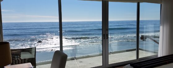 A serene coastal view from a modern bedroom with large floor-to-ceiling sliding glass doors, showcasing the glistening ocean waves and a clear blue sky. The room features a desk, a chair, and part of a bed in the foreground.