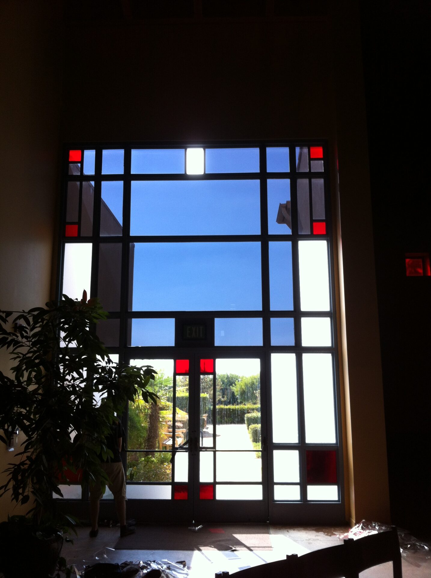 Large, modern glass window with a door at the bottom center. The window has a grid-like pattern with red and white accents. Sunlight shines through, illuminating the interior space. A plant is visible on the left side near the window.