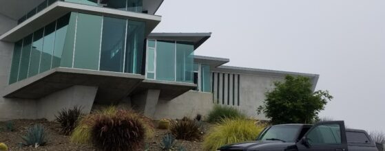 Modern, angular house with large glass windows sits on a raised hill, featuring minimalist landscaping with various grasses and shrubs. In the foreground, a black pickup truck is parked on a black asphalt driveway. The sky is overcast.