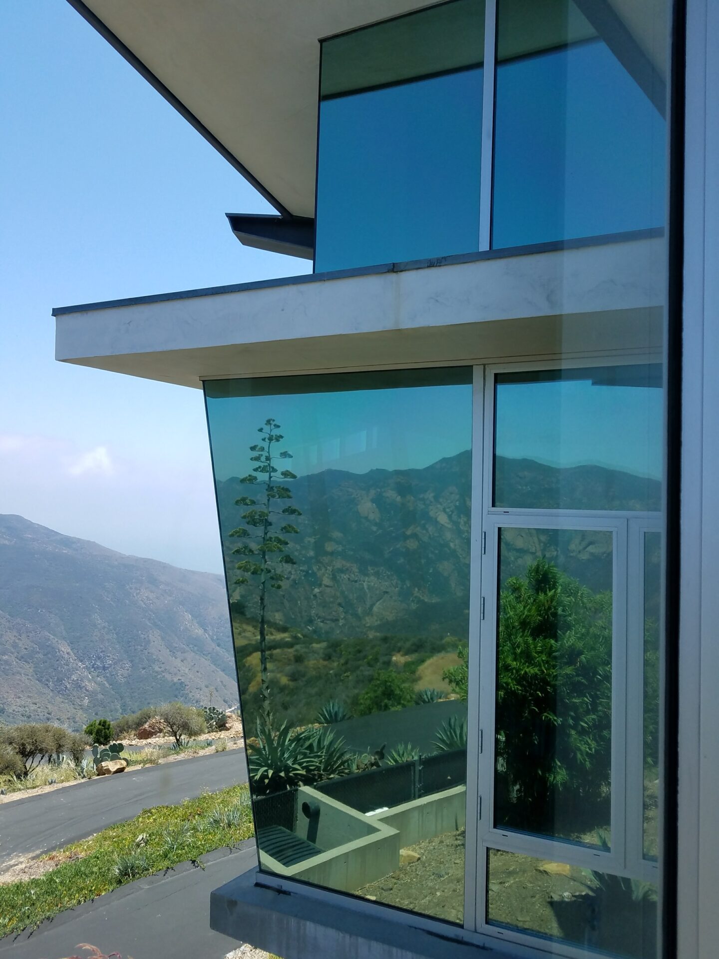 A modern glass-walled building with sharp, angular lines overlooking a scenic mountain landscape. The large windows reflect the mountainous horizon, blending the architecture with the natural surroundings. The sky is clear with a few scattered clouds.