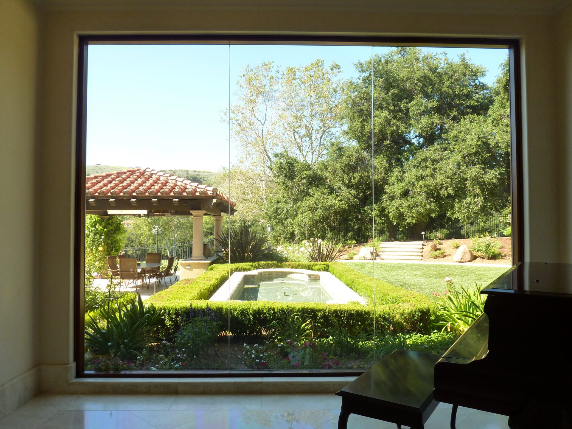 A large window offers a picturesque view of a well-manicured garden featuring a small pool surrounded by hedges. In the background, there's a charming gazebo with a tiled roof and outdoor furniture. Part of a grand piano is visible in the foreground.