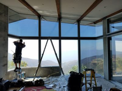 A person standing on a small ladder, installing or cleaning a large floor-to-ceiling window in a modern, unfinished room with wooden beams. A scenic mountain view is visible through the windows. Various tools and materials are scattered on the floor.