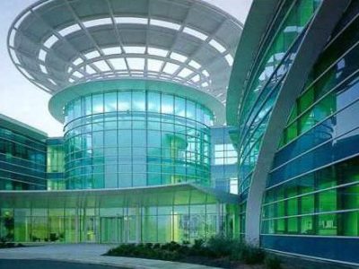 Modern glass building with a circular atrium and a unique dome-like structure on top, surrounded by curved, green-tinted glass windows. The design features sleek lines and reflective surfaces, complemented by landscaped greenery at the entrance.