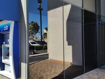 A photo showing two Citibank ATMs on the left side mounted on the wall of a building. The building features large reflective glass panels and a gated area on the right. A tree, lamp posts, and vehicles are visible in the reflection on the glass.