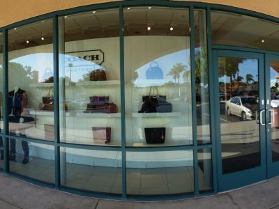 A wide-angle view of a Coach Factory store with glass windows and doors. Inside, various handbags are displayed on shelves. The store sign "Coach Factory" is visible above the entrance on the left. Reflections of the street and vehicles can be seen in the windows.