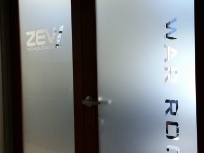 A set of double glass doors with wooden frames. The frosted glass has "ZEV Technologies" etched on the left door and "WAR ROOM" on the right door. There is a small, white sign above the doors with the word "WAR ROOM" written in black.