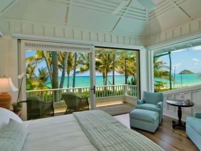 A cozy bedroom with white walls and light blue accents opens to a patio overlooking a tropical beach. Two armchairs and a small table are set near large sliding glass doors, providing a stunning view of palm trees and a turquoise ocean.