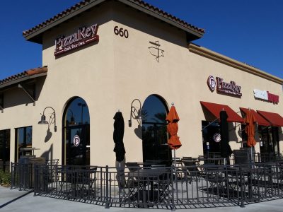 A street view of a pizzeria named "PizzaRev" with beige exterior walls and large arched windows. The patio area features tables shaded by orange and black umbrellas. The building is located at 660 and has other store signage visible.