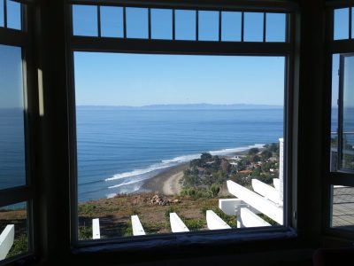 A breathtaking view of a calm, blue ocean framed by a large window. Below, a coastal village stretches along a curved shoreline with gentle waves lapping at the beach. Green hills and clear blue skies dominate the background, enhancing the serene atmosphere.